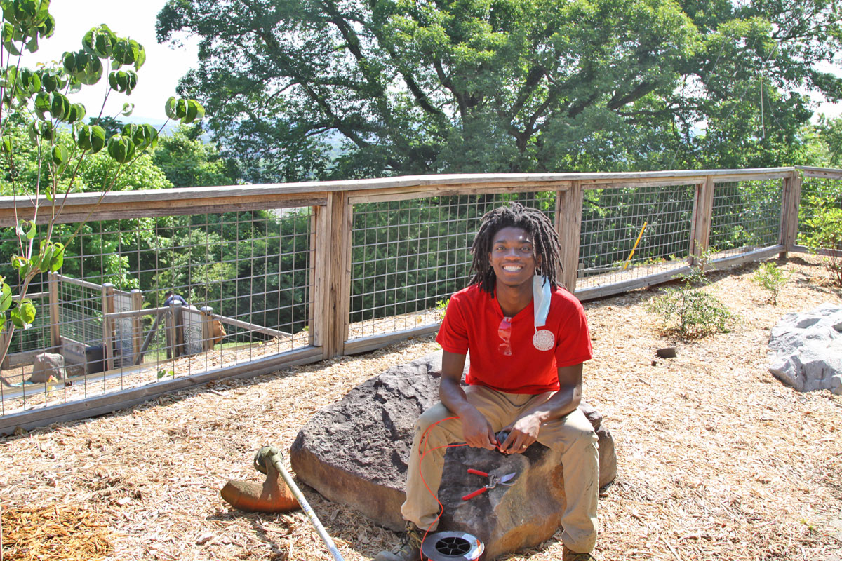 A young black man in a red shirt sits on a rock while changing the string in a weedeater.