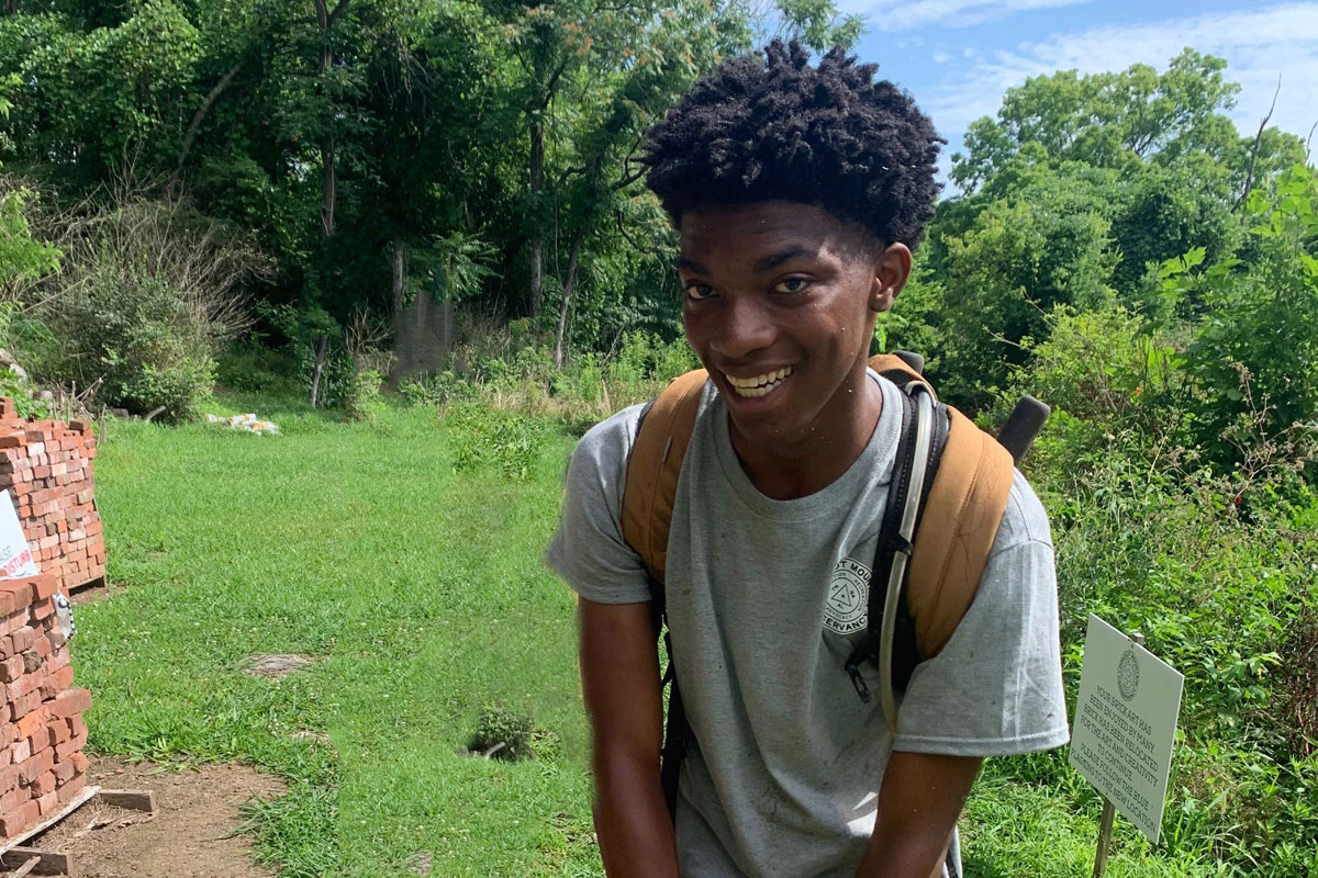 A young black man wearing a gray shirt and backpack smiles while working outdoors.