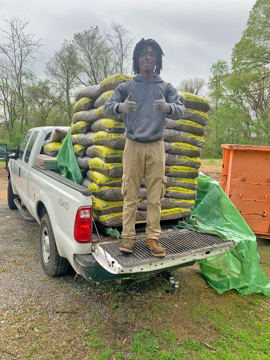 A young man stands in the bed of a pick-up filled with bags of mulch and gives two thumbs up.
