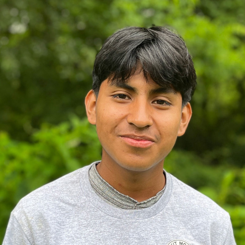 Headshot of a smiling young Hispanic male in a grey shirt before a forested background