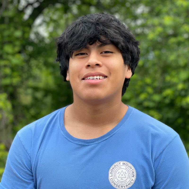 A young Latino male in a blue shirt poses with a smile in front of a forested background