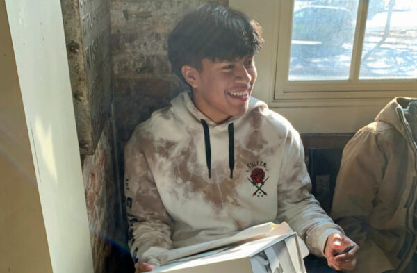 A smiling young Latino male sitting in front of a brick wall with a window