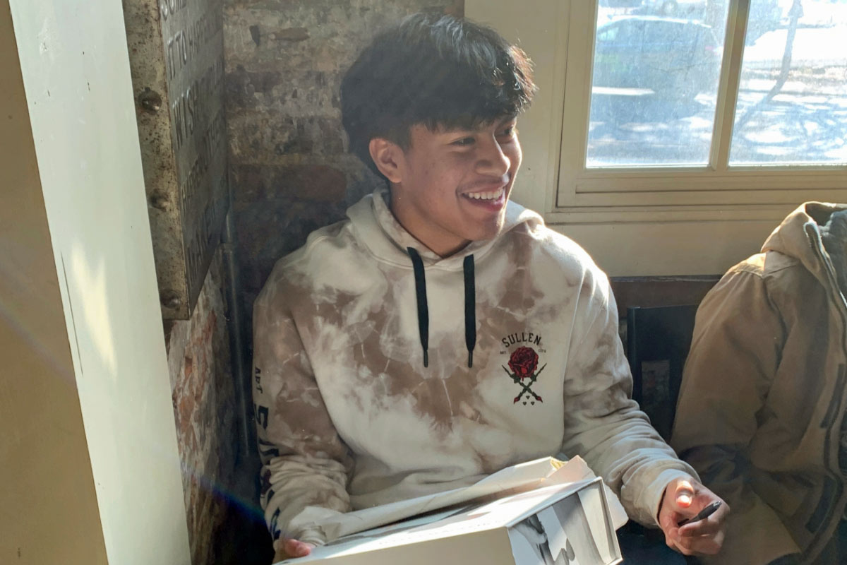 A smiling young Latino male sitting in front of a brick wall with a window