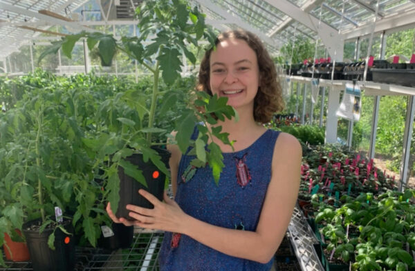 A young woman in a blue jumper with curly brown hair and white skin poses with a plant in a greenhouse