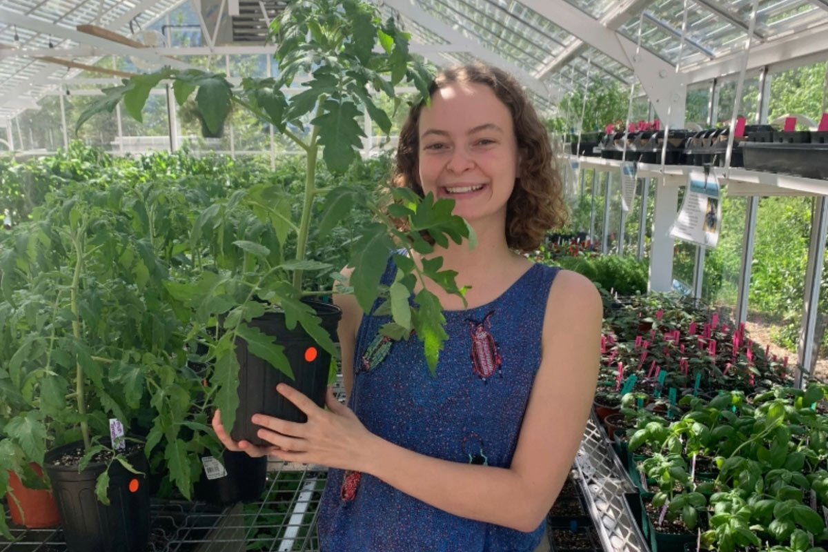 A young woman in a blue jumper with curly brown hair and white skin poses with a plant in a greenhouse