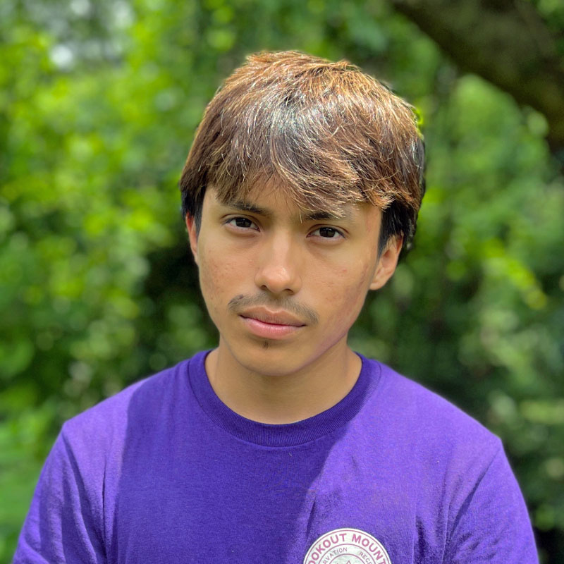 Headshot of a young man with dark hair wearing a purple shirt standing in front of a forested background