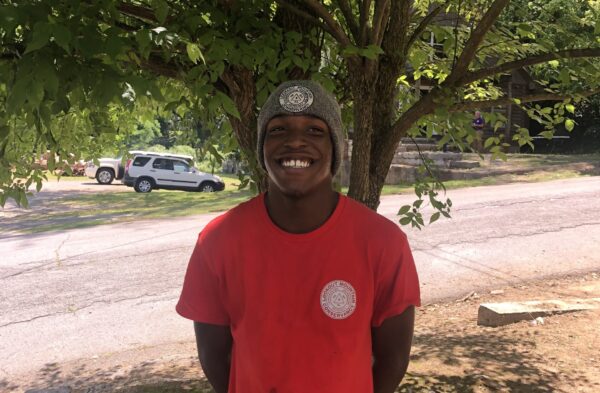 young black male teen smiling in a red shirt and grey beanie in front of a tree