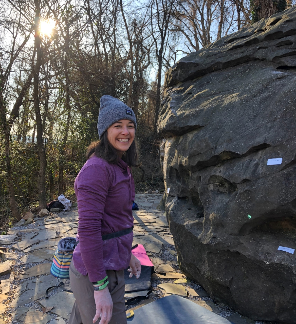 A smiling woman poses in front of a boulder she is about to climb.
