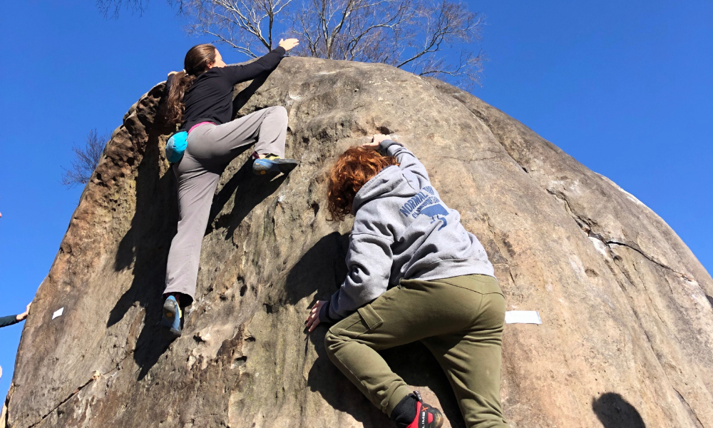 Two young people climb a boulder.