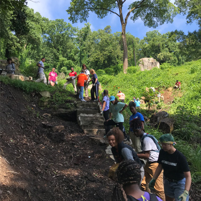 Interns and youth works forming a line to pass bags of dirt, sand, and gravel to the base of a boulder