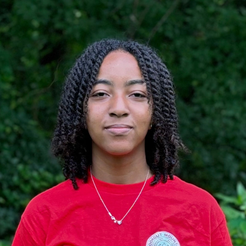Headshot of a young black woman with shoulder length curly black hair in a red shirt in front of a forested background