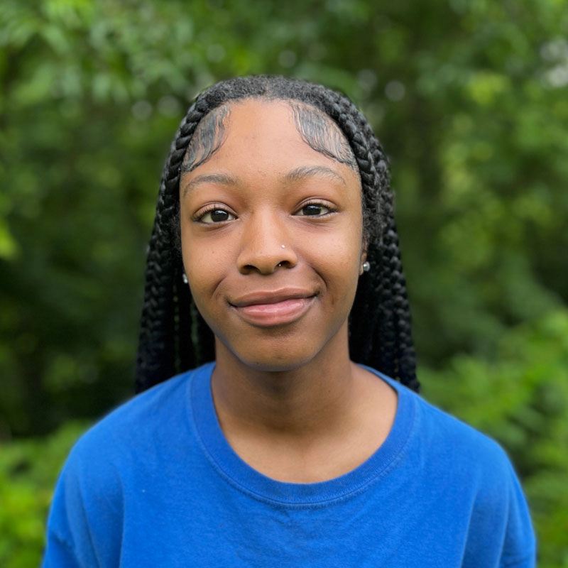 Headshot of a young black woman with long braided hair in a blue shirt in front of a forested background