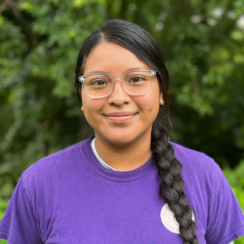 Headshot of a young woman with black braided hair and glasses in a purple shirt with a forested background