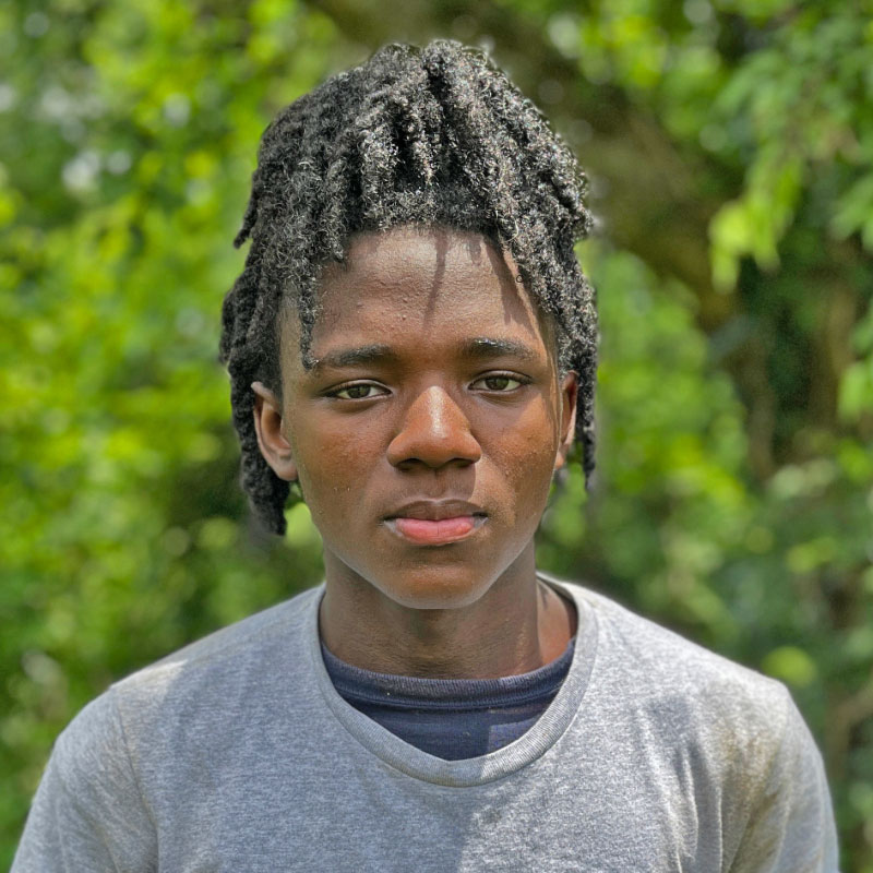 Headshot of a black teenage male with locks and a grey shirt in front of a forested background