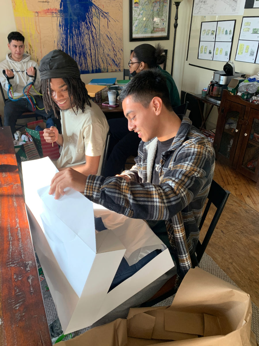 A smiling teenaged Latino male in a flannel shirt opens a gift next to another young man.