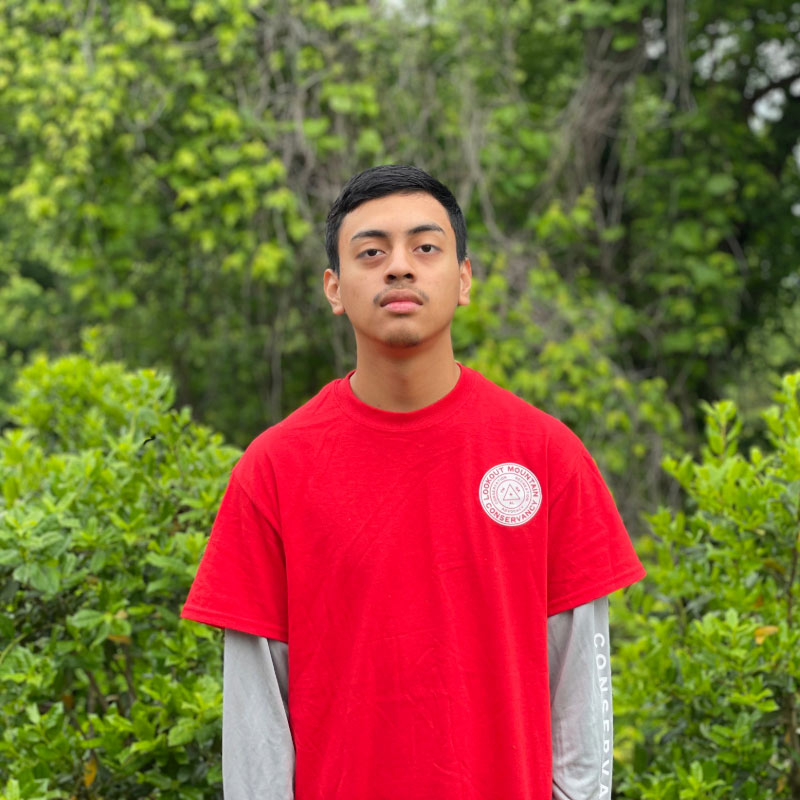 A male Latino teenager in a bright red shirt poses in front of a forested background.