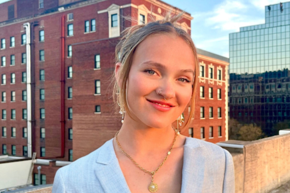 Headshot of a young white female in an urban setting with blond hair and wearing a light grey suit.