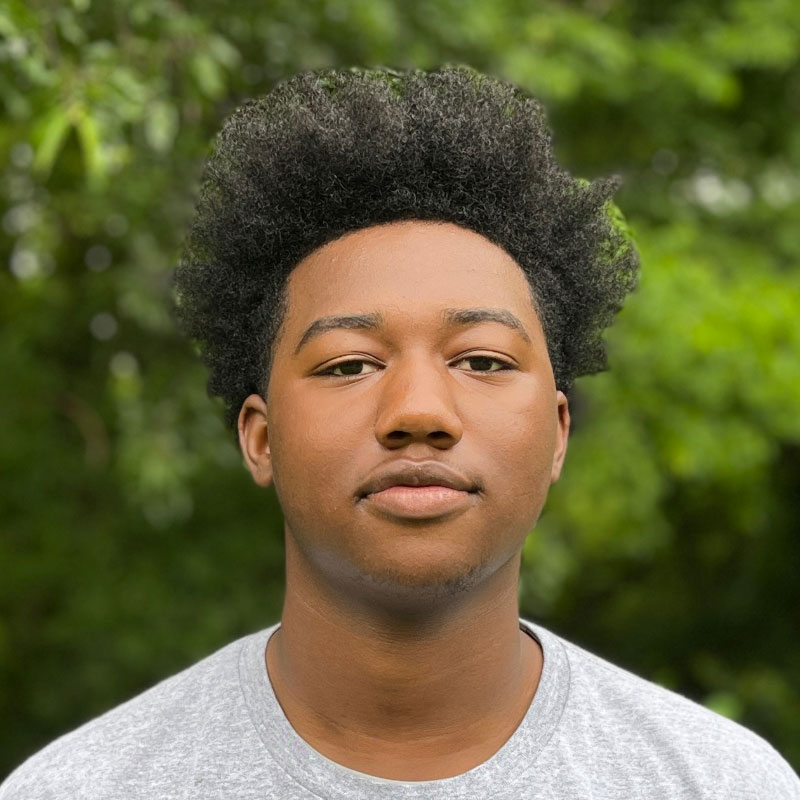 Headshot of a young black male in a grey shirt with a short afro in front of a forested background