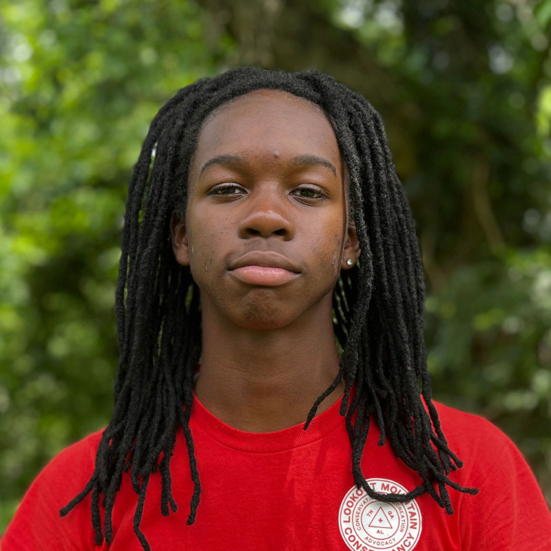 Headshot of a black male teenager with long locks in a red shirt before a forested background