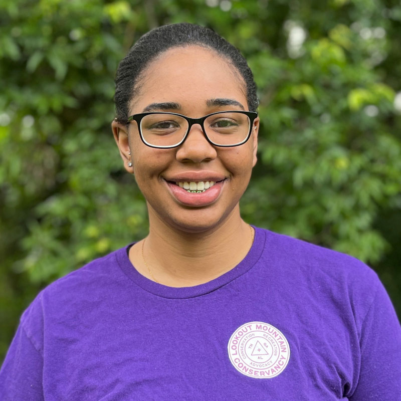 Headshot of a young black woman with black glasses and a purple shirt in front of a forested background