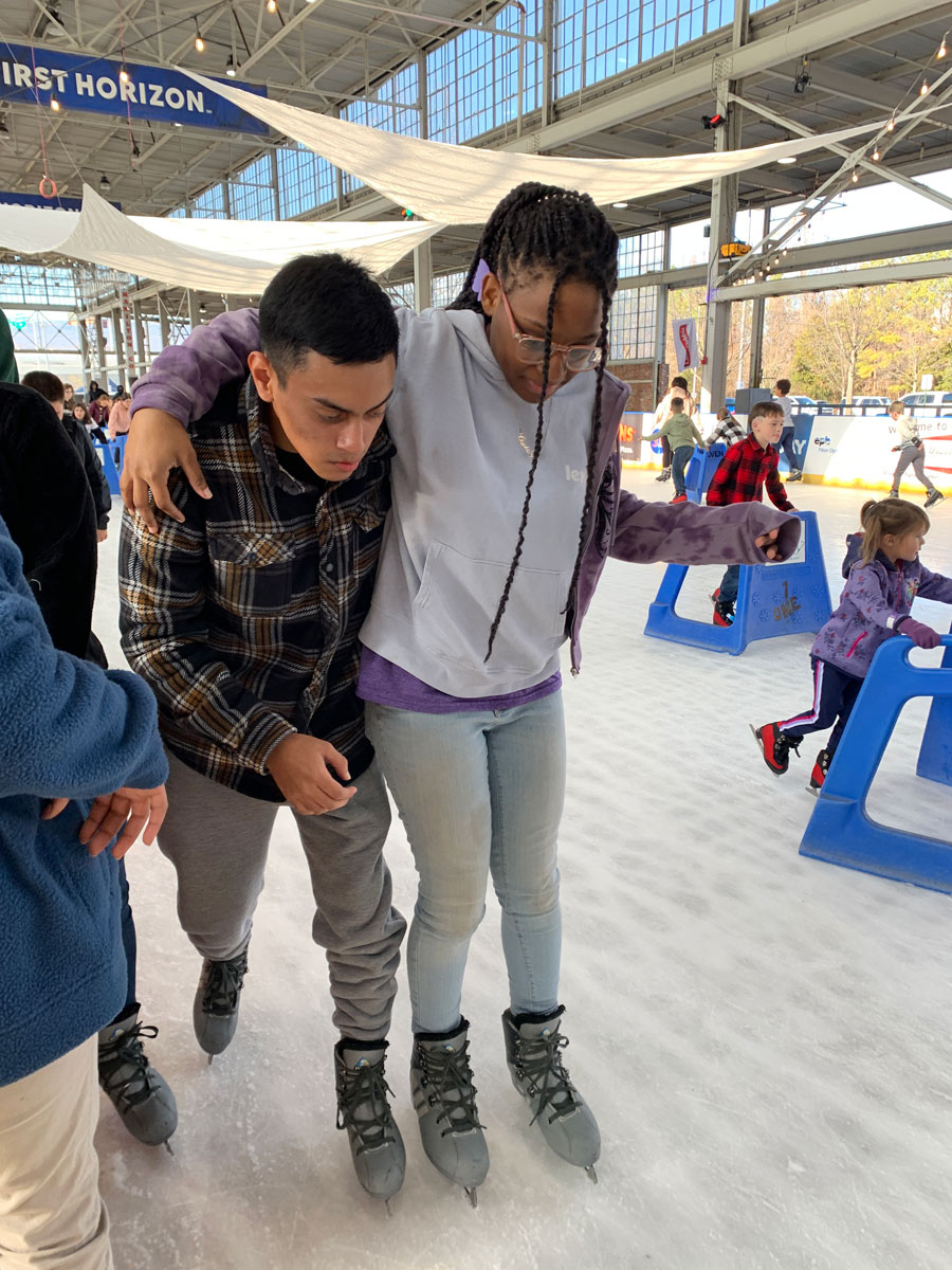 Two teenagers ice skate side by side. A black female with her arm over the shoulder of a Latino male.