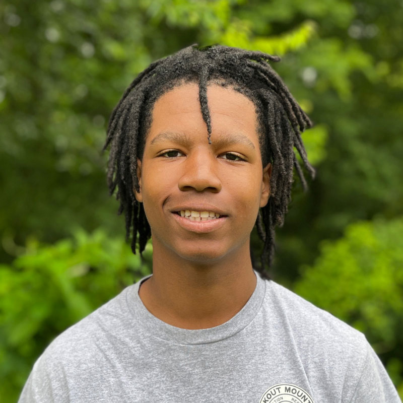 Headshot of a young black male with locks and a grey shirt in front of a forested background