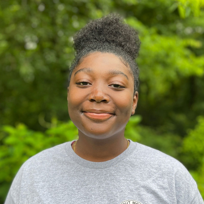 Headshot of a smiling black teenage girl with her hair in a bun, wearing a grey shirt in front of a forested background