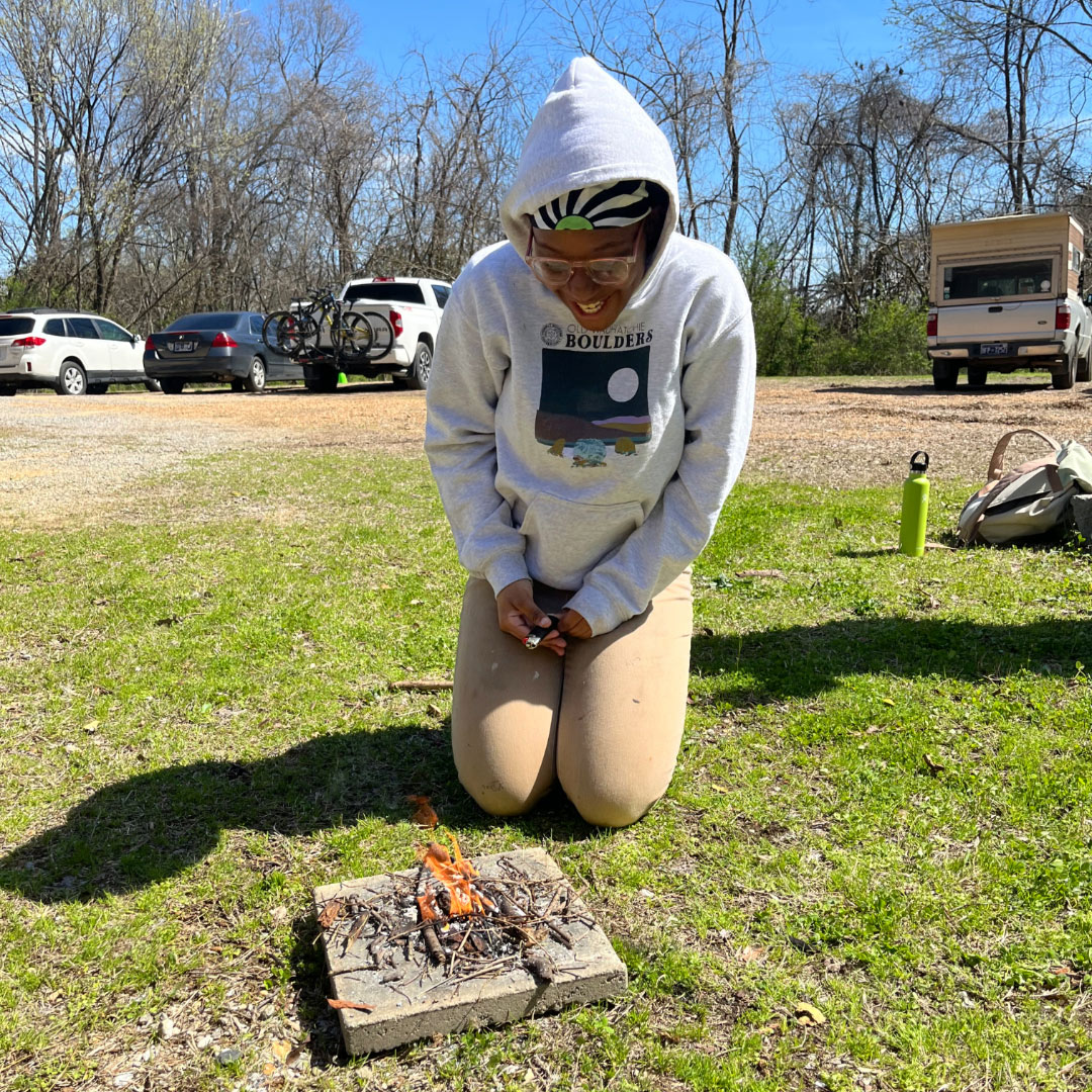 A young woman in a gray hoodie smiles over the beginnings of a small fire she started.