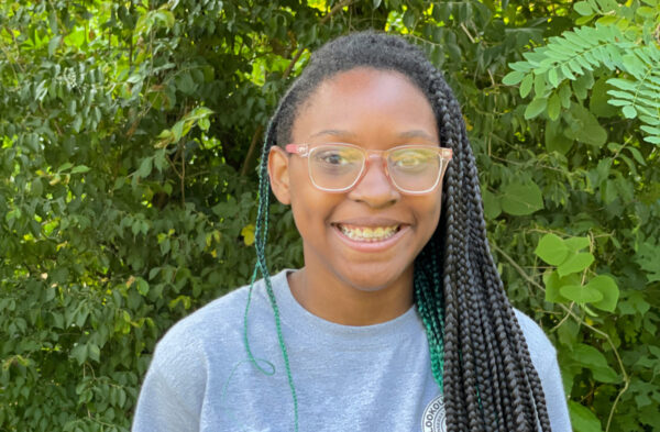 A young black woman with glasses and braids before a forested background.