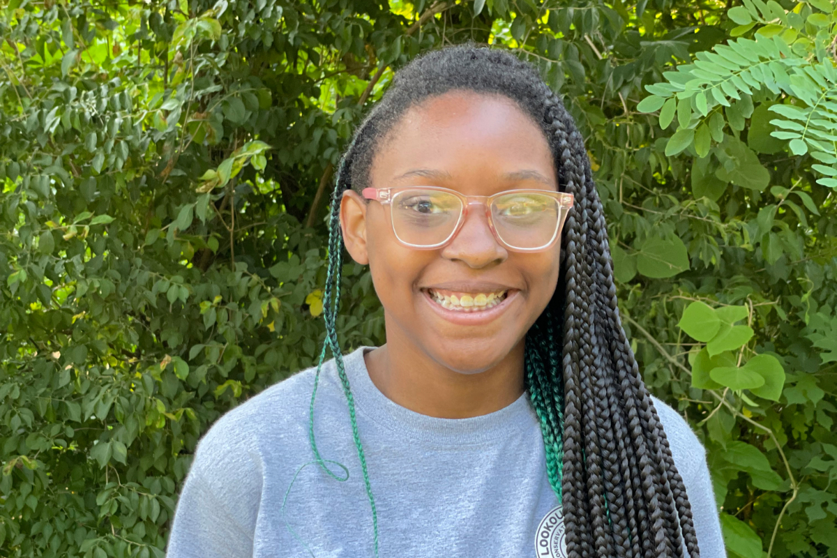 A young black woman with glasses and braids before a forested background.