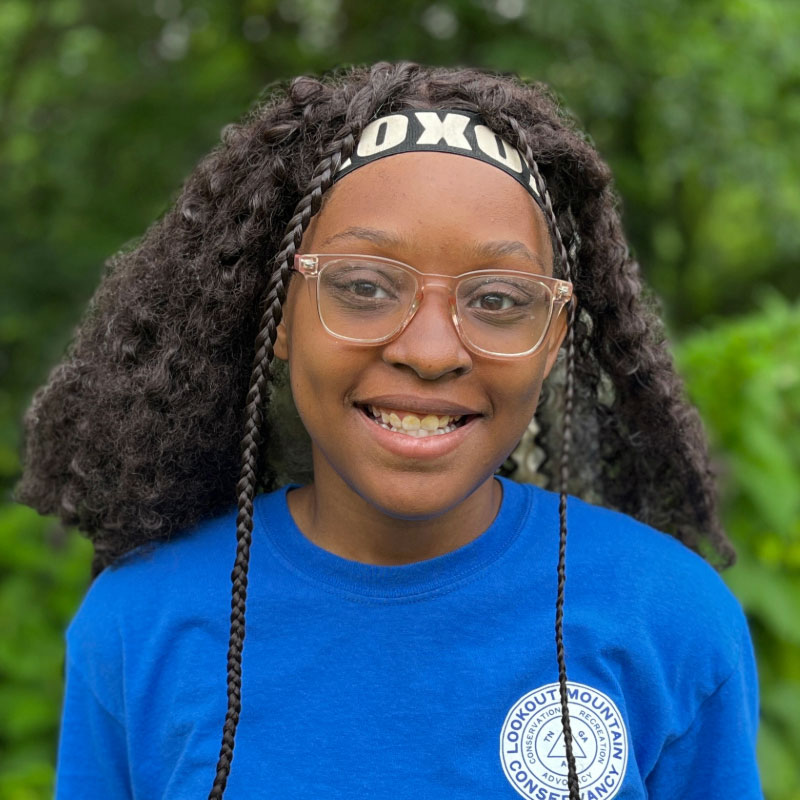 Young black woman with curly black hair in clear glasses and a blue shirt in front of a forested background