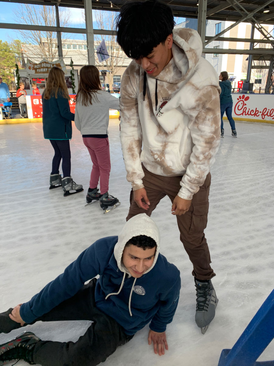 A young man leans over to help another young man stand back up after falling while ice skating.