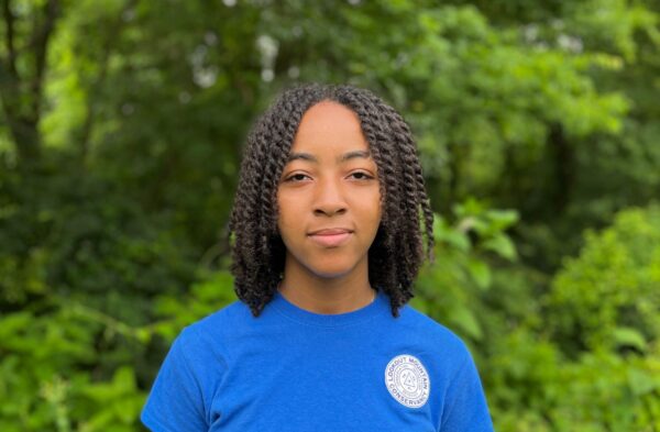 Young black female smiling in a blue shirt in front of a forest backdrop.
