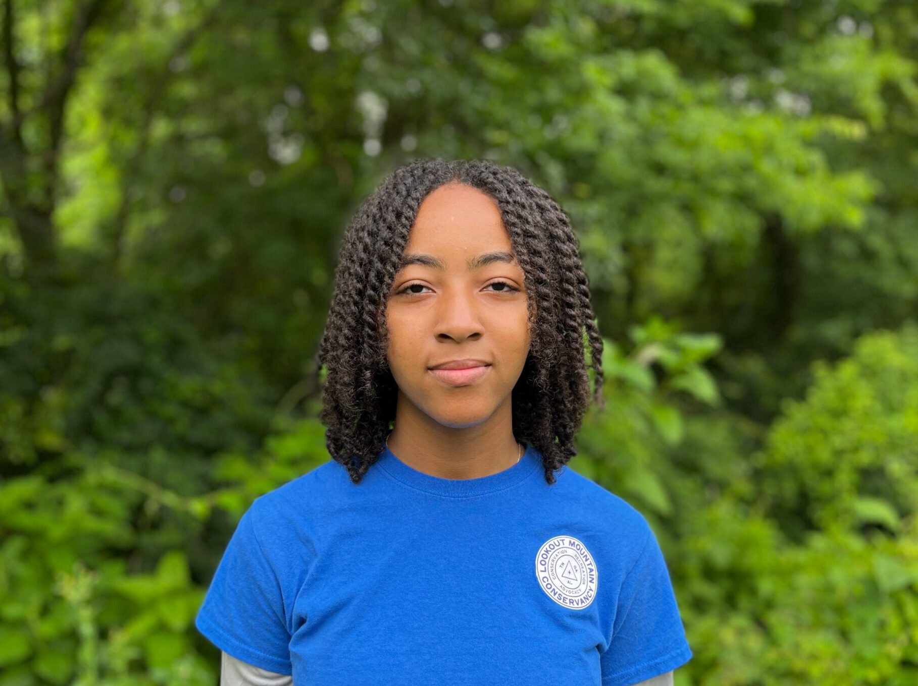 Jada Heazdshot Young black female smiling in a blue shirt in front of a forest backdrop.