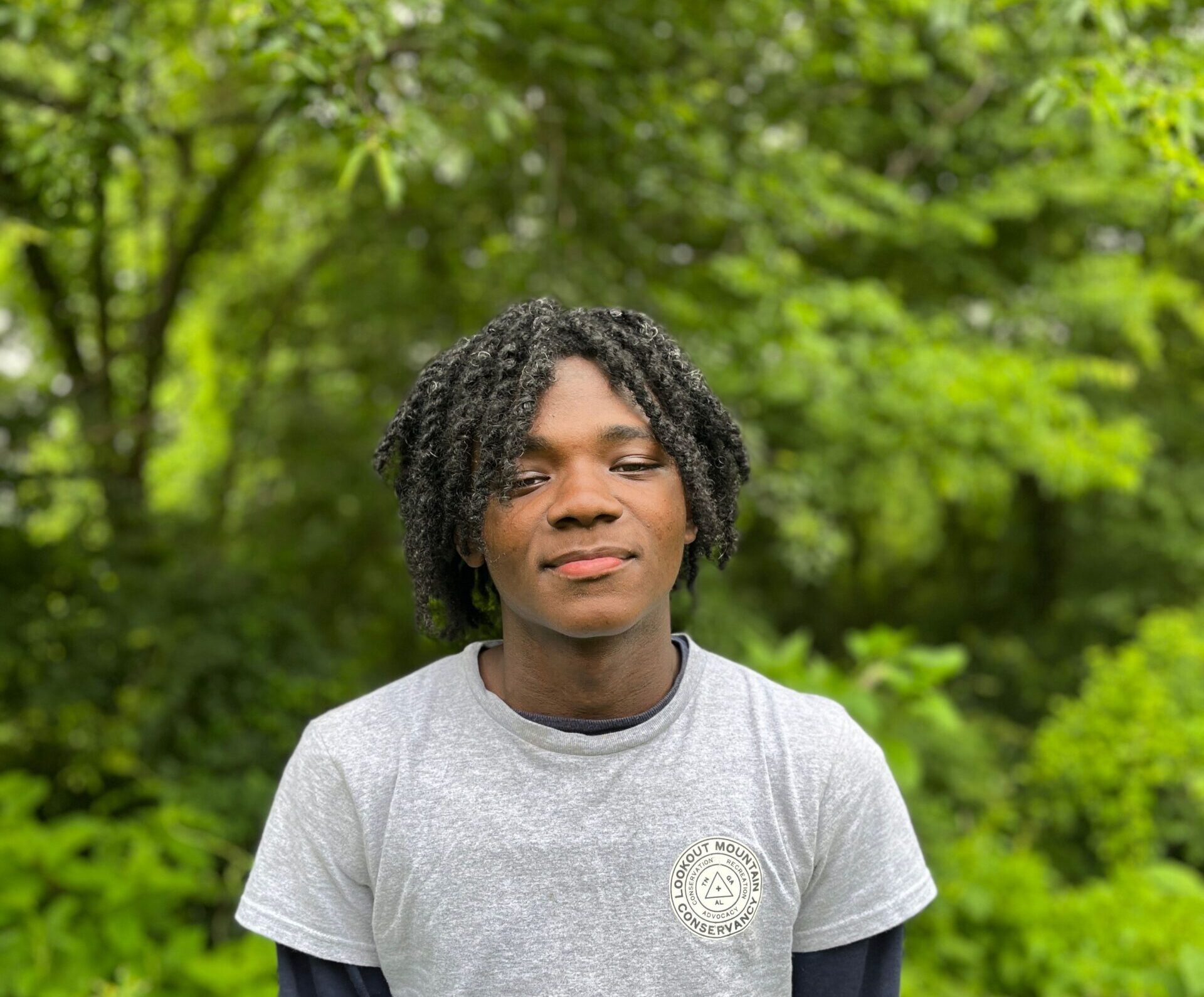 John Headshot young black male smiling in a grey shirt in front of a forest.