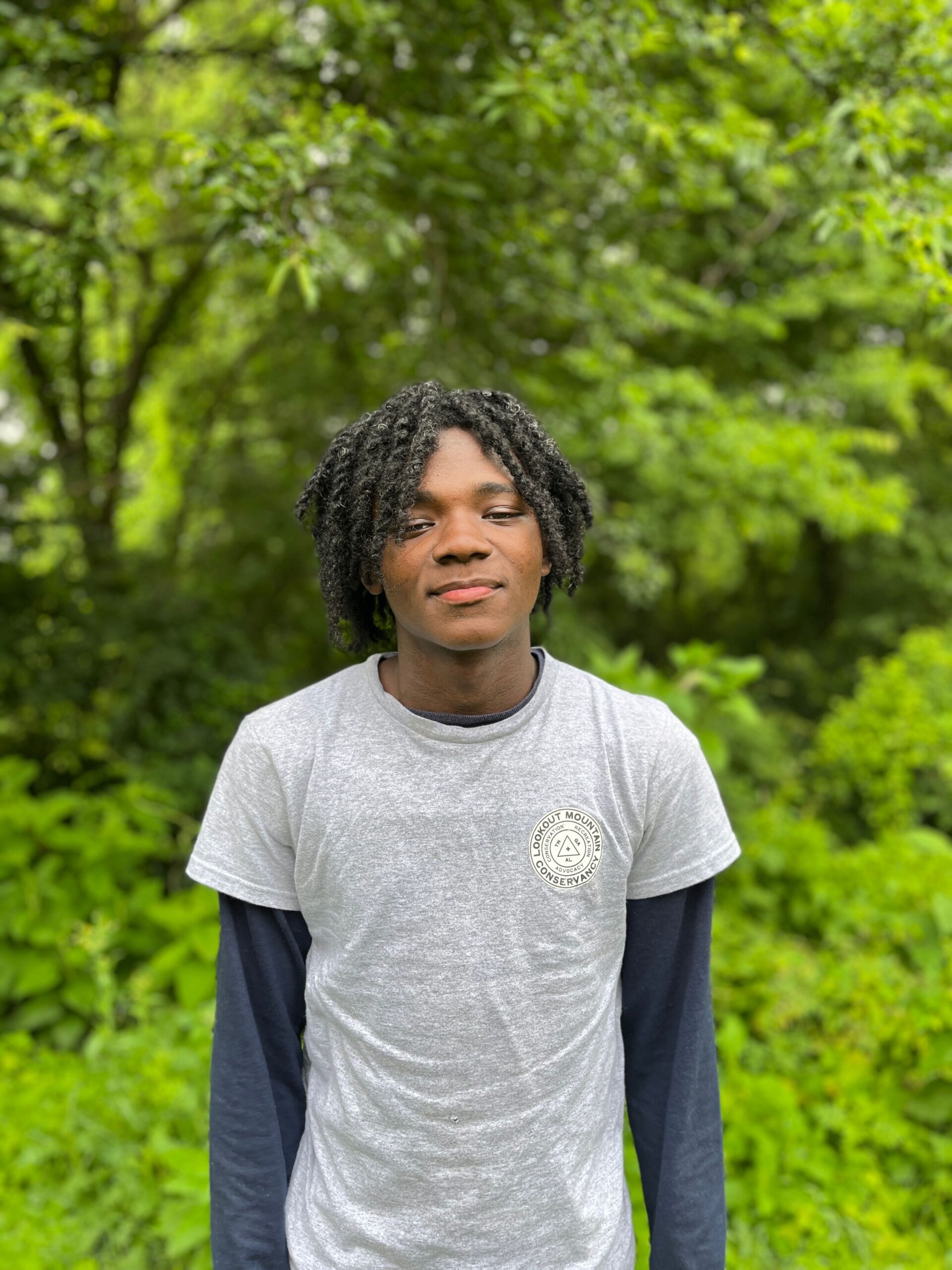 young black male smiling in a grey shirt in front of a forest.