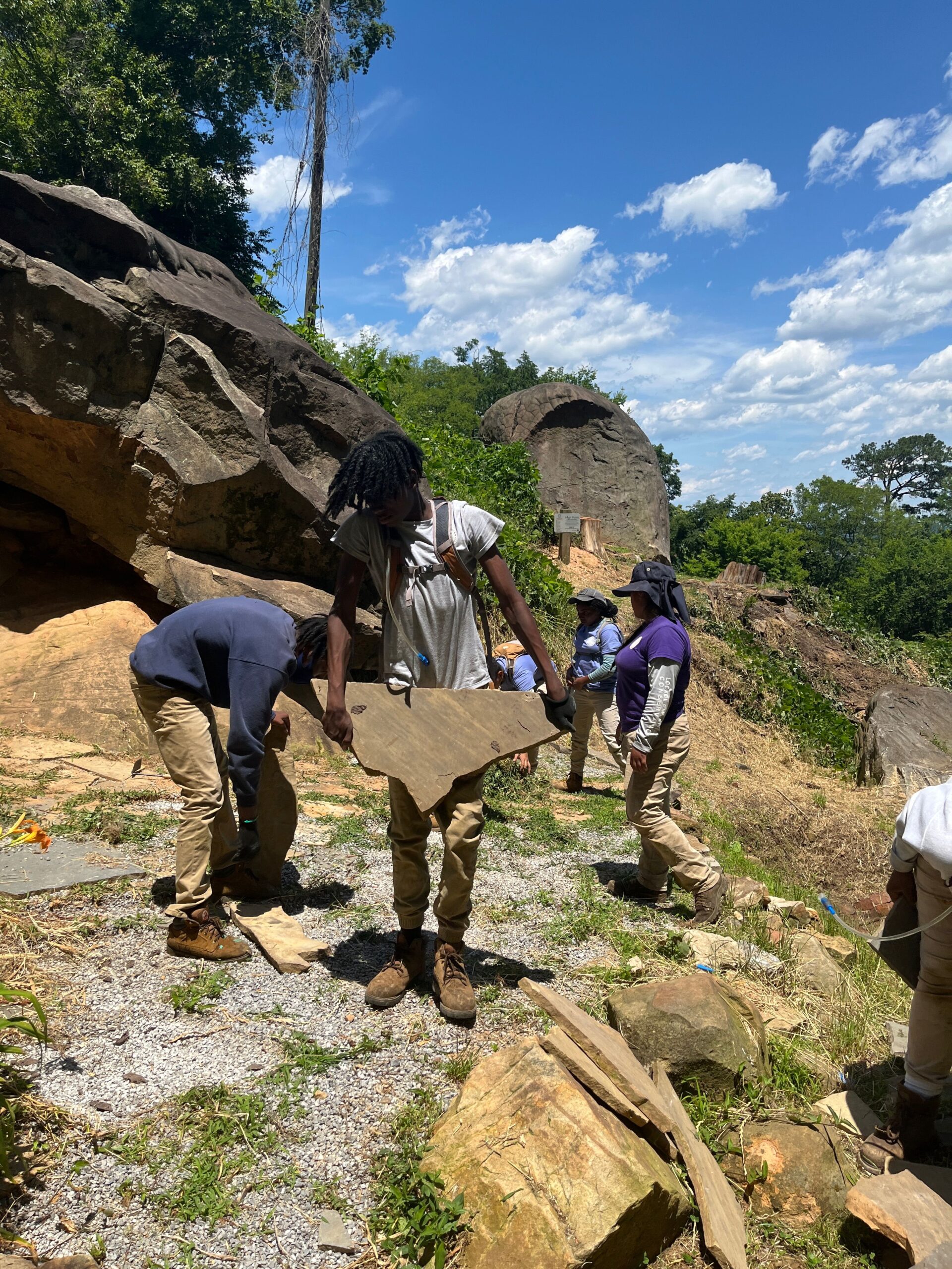 young black male in a grey shirt carrying stone on a work site.
